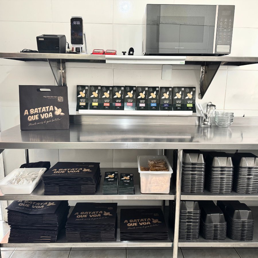 Bancada inox da segunda cozinha com embalagens A Batata Que Voa, utensílios e bandejas empilhadas.