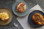 Três opções de batata recheada servidas em pratos sobre mesa escura, com recheios variados e visual artesanal.