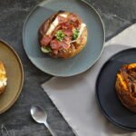 Três opções de batata recheada servidas em pratos sobre mesa escura, com recheios variados e visual artesanal.