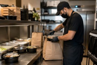Operador embala pedido em dark kitchen moderna com balcão inox, marmitas prontas e bolsas de delivery ao fundo.