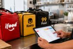 Gestor usa tablet na cozinha com bolsas de apps de delivery sobre bancada de inox iluminada pela manhã