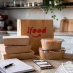 Sacola do iFood e embalagens de delivery sobre bancada de cozinha, com celular e caderno para vender no iFood.