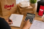 Cálculo de quanto sobra no iFood, com pedidos, calculadora, celular e anotações de custos na mesa.