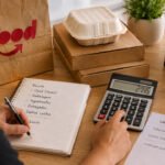 Cálculo de quanto sobra no iFood, com pedidos, calculadora, celular e anotações de custos na mesa.